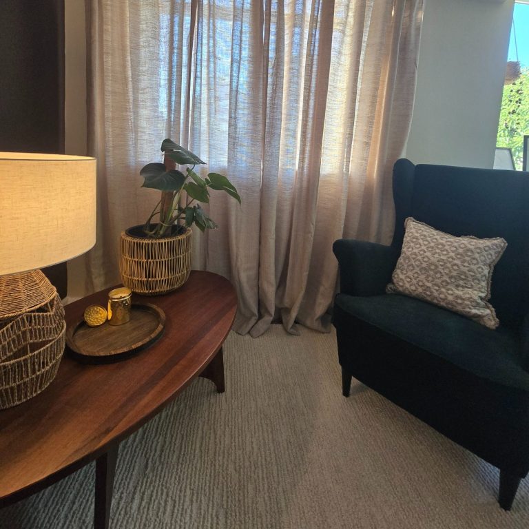 A cosy room with warm lighting. A green wingback armchair with cushion are on the right. A plant, candle and lamp are set on a table.