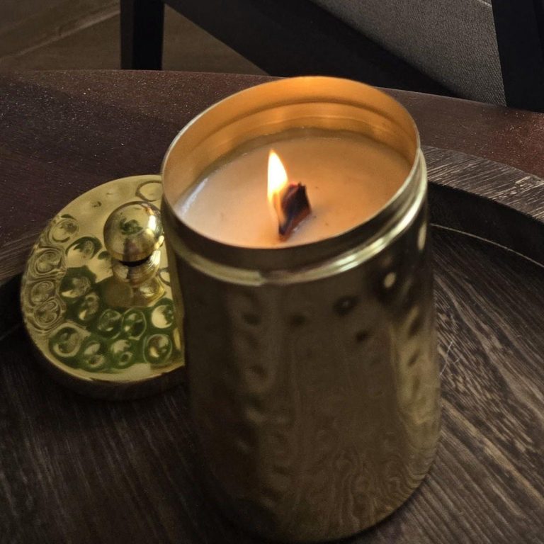 A cream coloured candle in a gold candle holder with a wooden wick and flame 