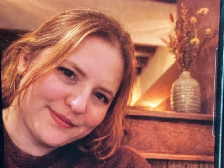 Photo of a Caucasian female with light brown/blonde hair smiling. Wearing a brown jumper. A vase with dried flowers is in the background 